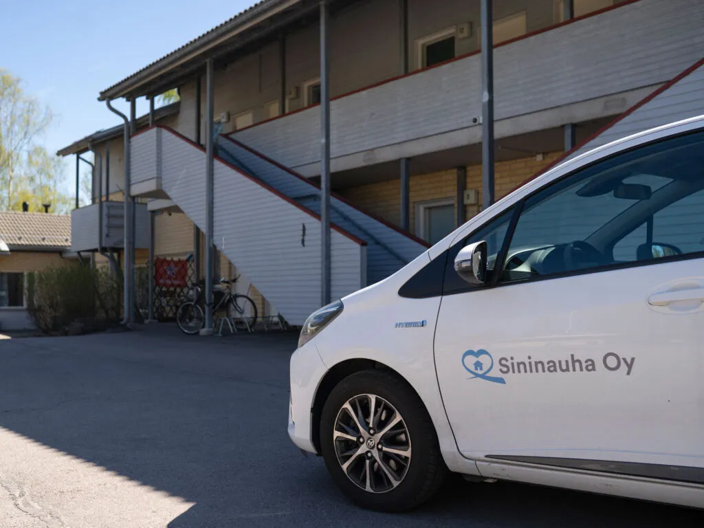 Sininauha Oy:n logolla varusteltu auto parkkeerattuna keltatiilisen luhtitalon pihaan.
