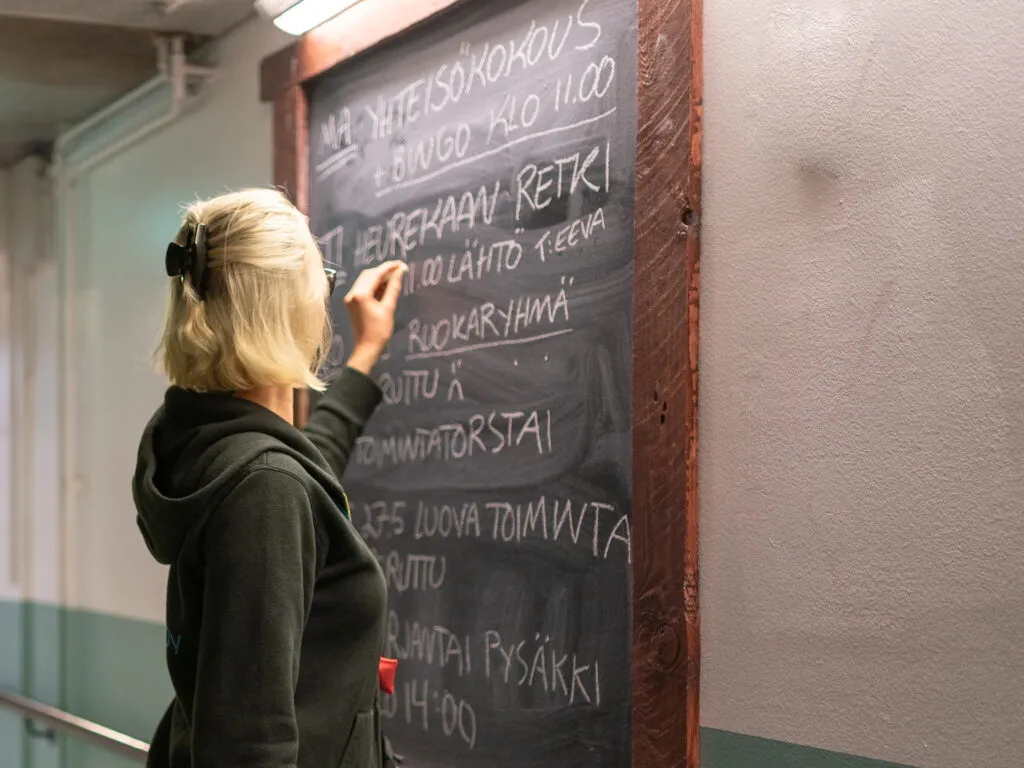 Henkilö kirjoittaa rappukäytävässä liitutaululle viikon ohjelmaa, johon kuuluu muun muassa retki Heurekaan, ruokaryhmä ja luovaa toimintaa.