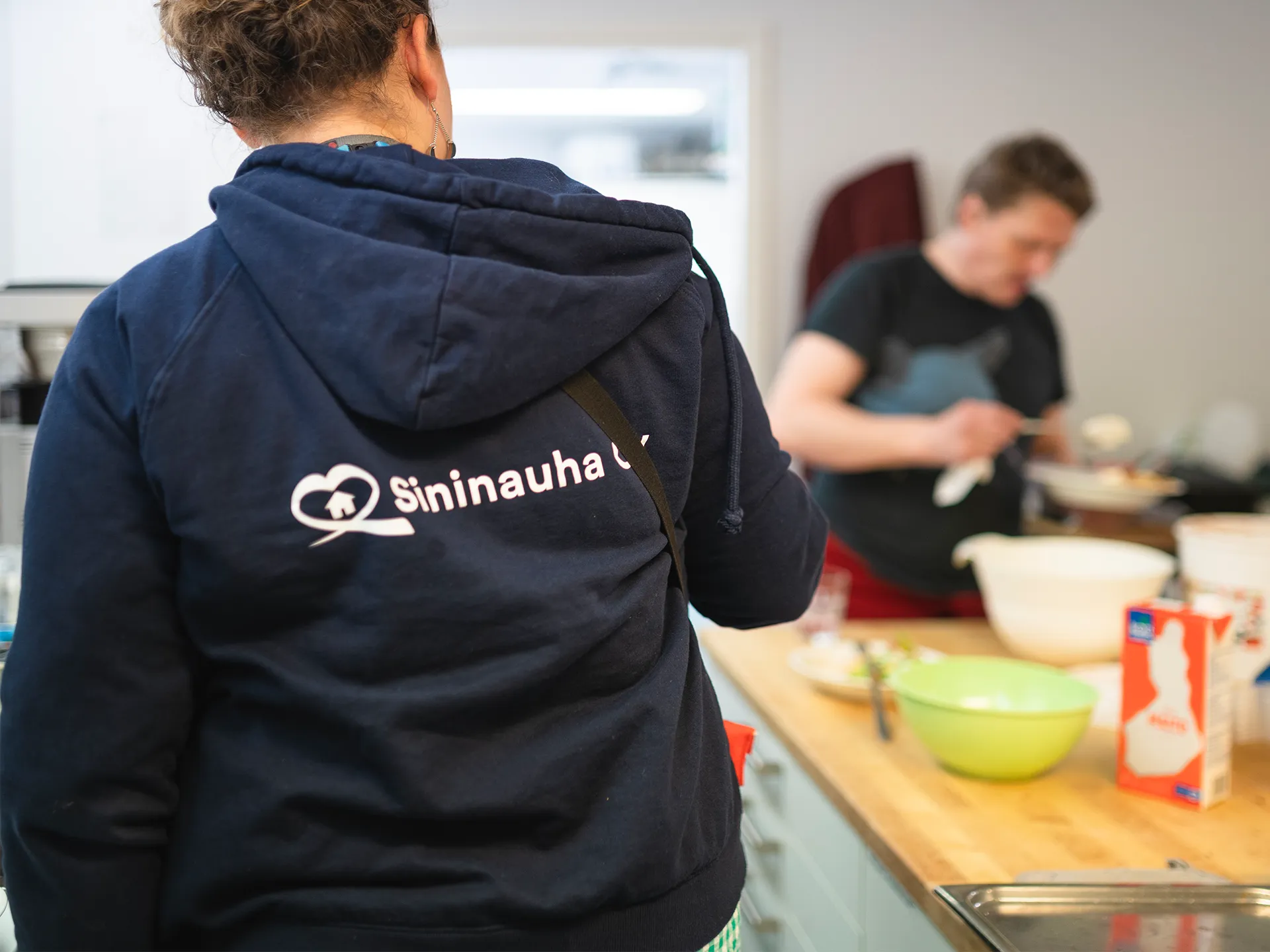 Kamera on tarkentunut työntekijän huppariin, jonka selässä lukee Sininauha Oy. Taustalla henkilö ottaa ruokaa lautaselleen keittiösaarekkeelta.